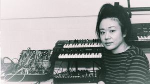 Black-and-white photo of Midori Hirano in a striped sweater, hair piled high, looking into the camera beside stacked synthesizer keyboards and a modular synth tangled with cables.