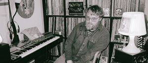 Sam Cook-Parrott in tinted glasses sits slouched between a keyboard and a white table lamp, record shelves and a Radiator Hospital sign behind him. Black and white.