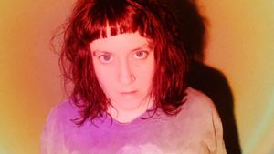 Tiiva in close-up, dark fringe and a nose ring, looking straight at the camera against a warm orange glow, a hard shadow cast behind them.