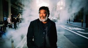Wadada Leo Smith stands on a city street with his hands in his jacket pockets and smiles slightly at the camera as steam drifts around him and pedestrians pass.
