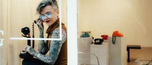 Through a storefront window, Whitney George, in tinted glasses, holds a vintage candlestick phone to one ear; colored rotary phones on plinths fill the room behind.
