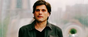 Brett Bullion of deep glens faces the camera in a dark jacket, city buildings blurred behind him through a misted window.