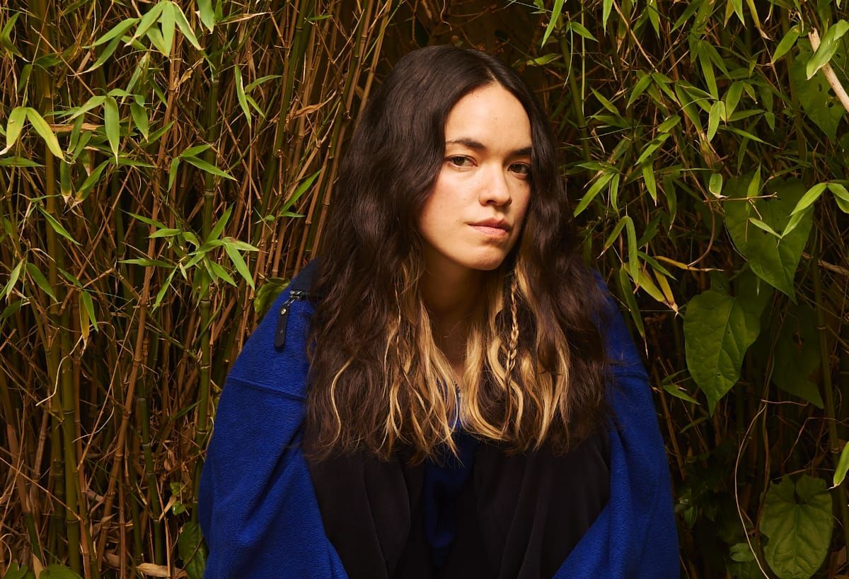 Momoko Gill with long wavy hair against bamboo backdrop, wearing blue jacket and black clothing in natural outdoor setting. Photo by Manuel Vasquez.