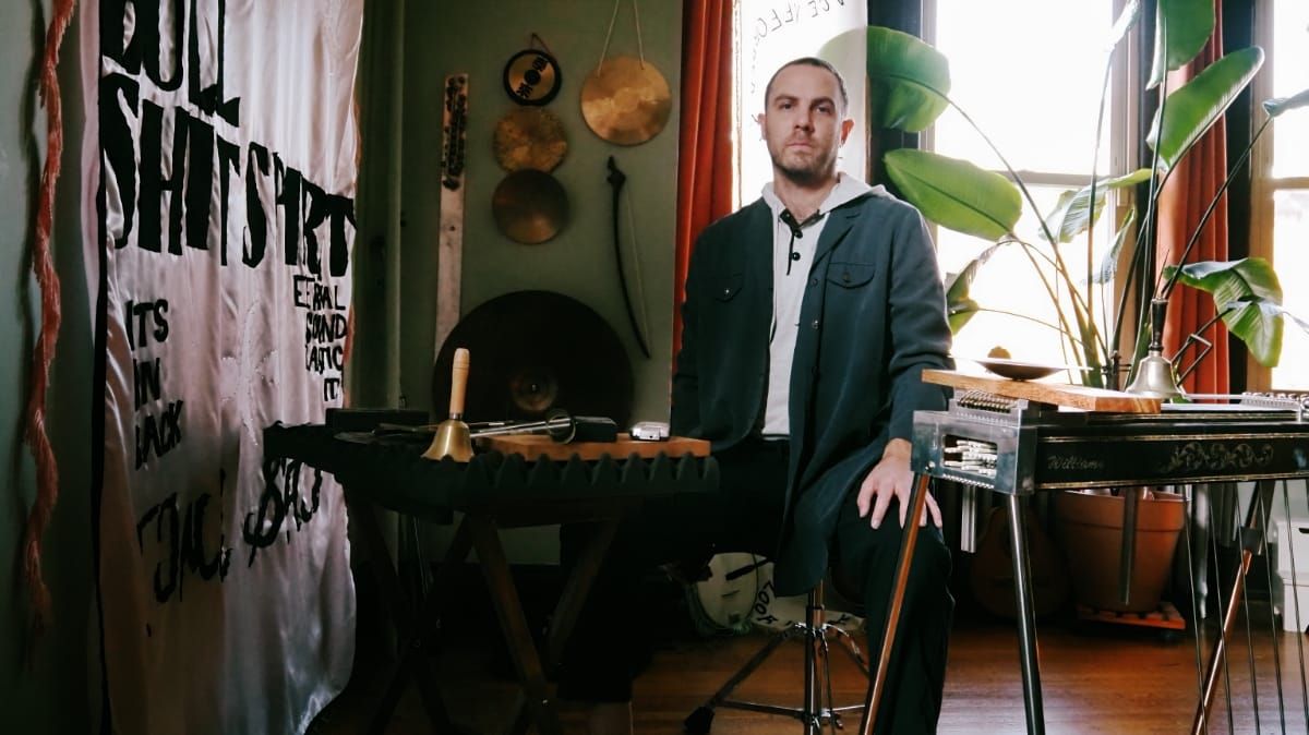 Sam Wenc in teal henley stands in home studio with pedal steel guitar, plants, hanging metalwork, and text banner, warm lighting.