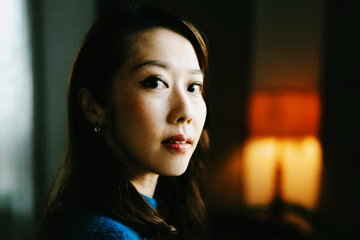 A close portrait of Miho Hazama in a blue top, lit from the side with a warm glowing lamp blurred in the background.