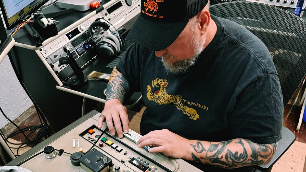 Billy Polo bends over a vintage tape machine console, both hands on the controls, in a studio with rack-mounted gear and headphones visible behind him.