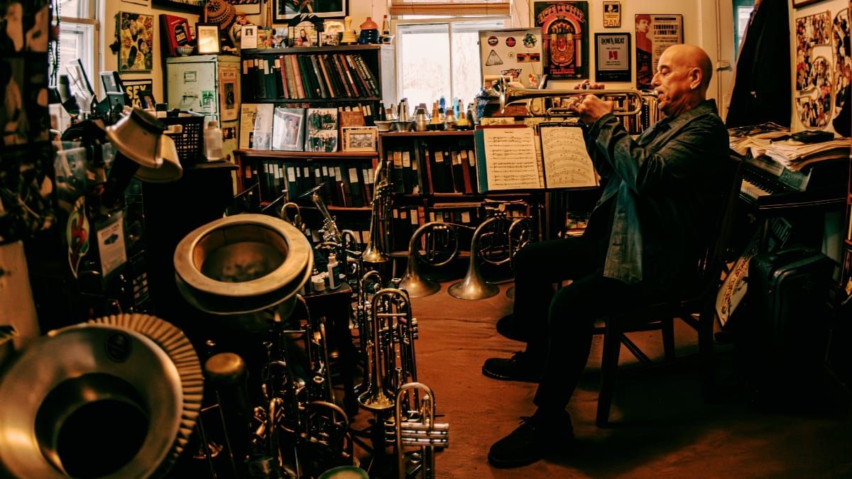 Steven Bernstein sits playing trumpet at a music stand in a cluttered studio crowded with brass instruments, horns, bookshelves, and posters lining the walls.