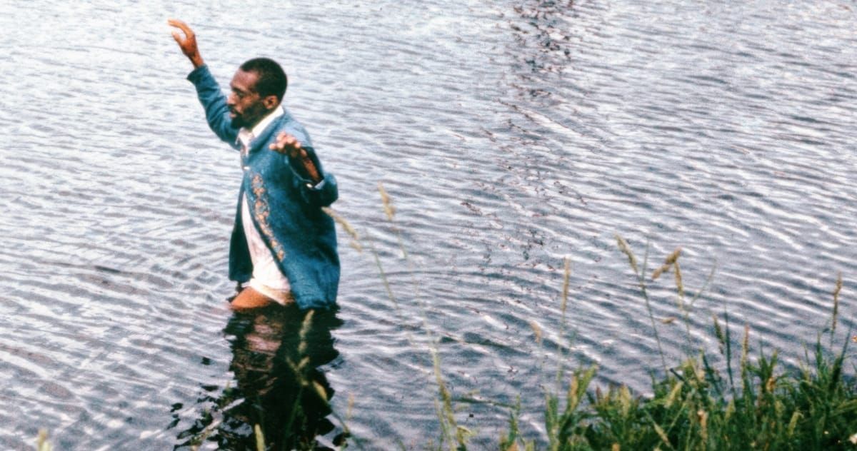 Julius Eastman stands waist-deep in a lake, one arm raised, the other extended outward, wearing a blue embroidered jacket over a white shirt.