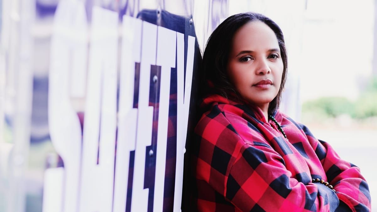 Terri Lyne Carrington leans against a window in a red and black plaid hoodie, arms folded, looking directly at the camera past the word SAFETY in large white letters on the glass.