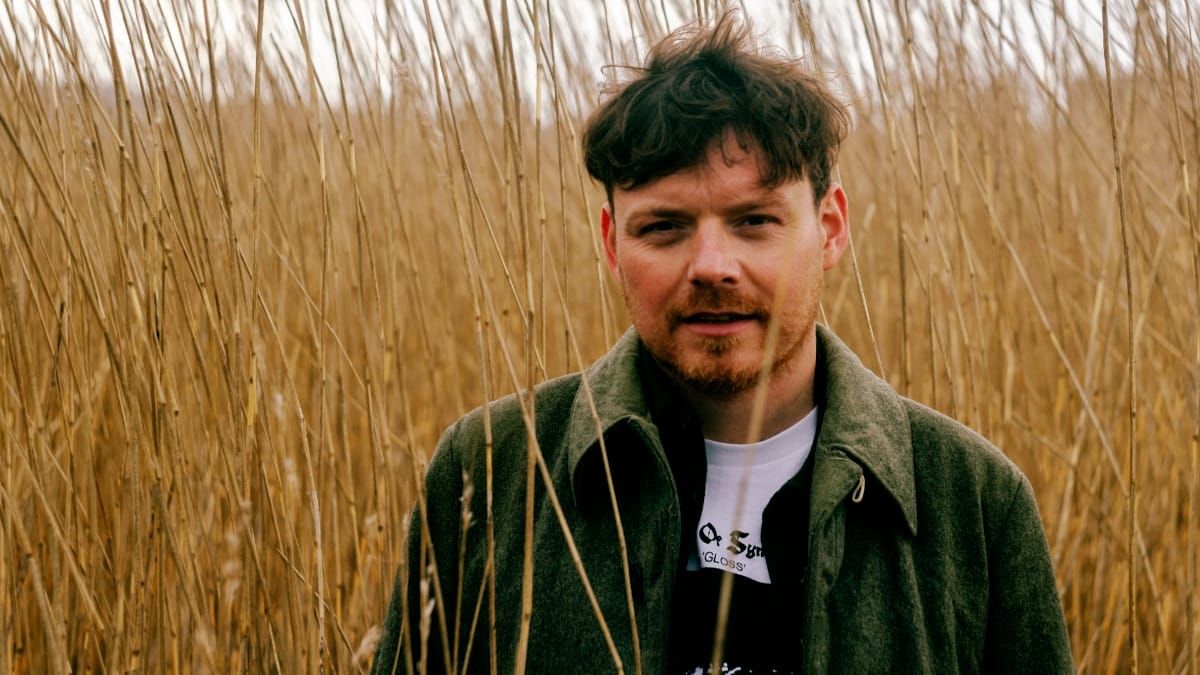 Jamie Livingstone stands among tall pale reeds that rise above his head, looking directly at the camera in an olive jacket over a white T-shirt.