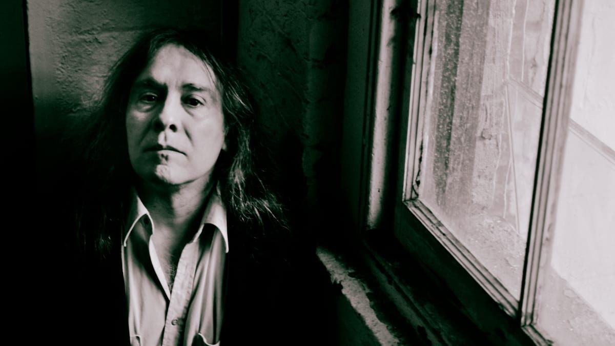 Martin Bisi leans against a wall beside a grimy window, looking into the camera, in a dark jacket over an open-collared shirt. Black and white.