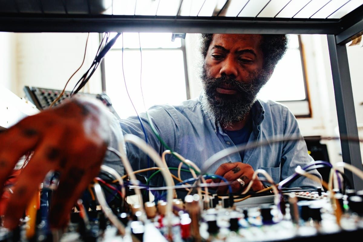 Robert Aiki Aubrey Lowe looks down at a modular synthesizer, framed by its patch cables and knobs in the foreground, window light behind him, a blurred hand reaching in from the left.