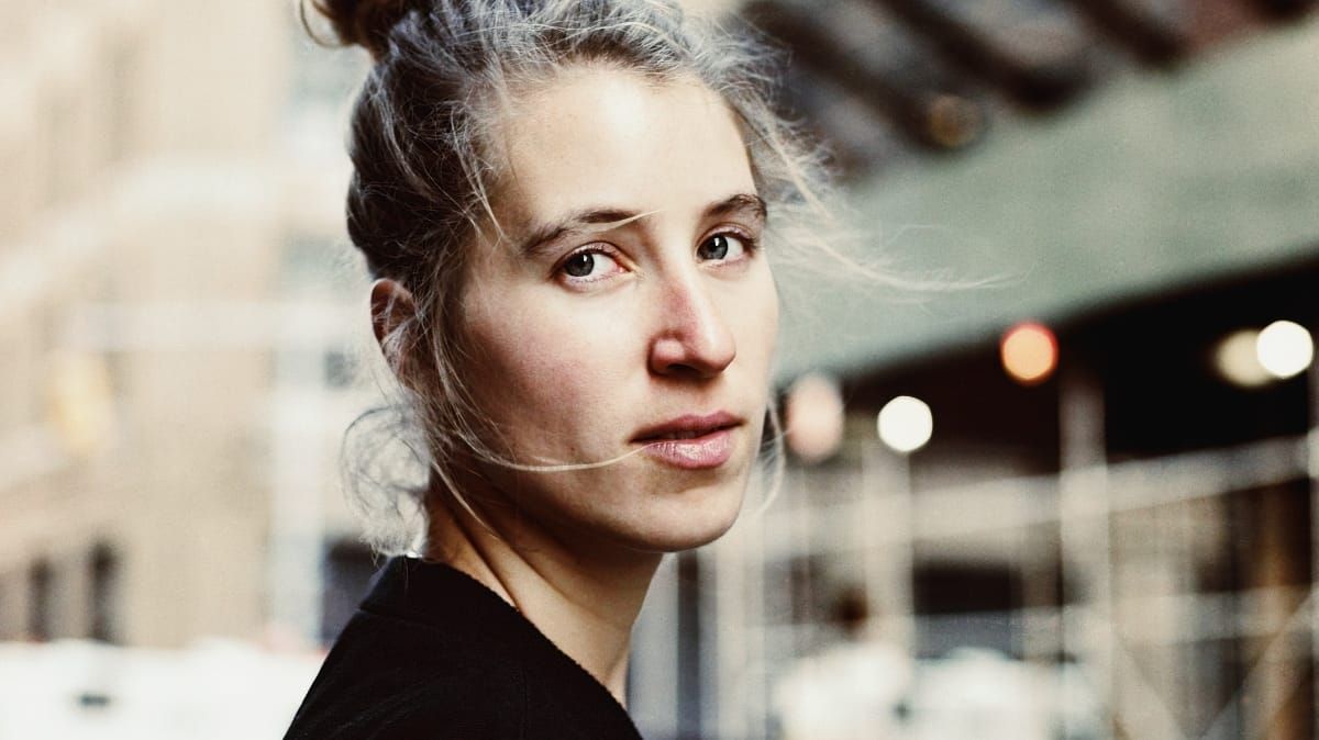 Charlotte Greve looks back over her shoulder at the camera, wisps of hair across her face, on a blurred city street.