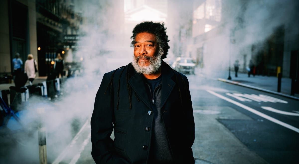 Wadada Leo Smith stands on a city street with his hands in his jacket pockets and smiles slightly at the camera as steam drifts around him and pedestrians pass.