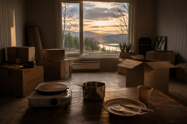 A sparsely furnished room filled with moving boxes, a small hotplate and a saucepan on a table, with evening light shining through a large window overlooking a quiet rural landscape.