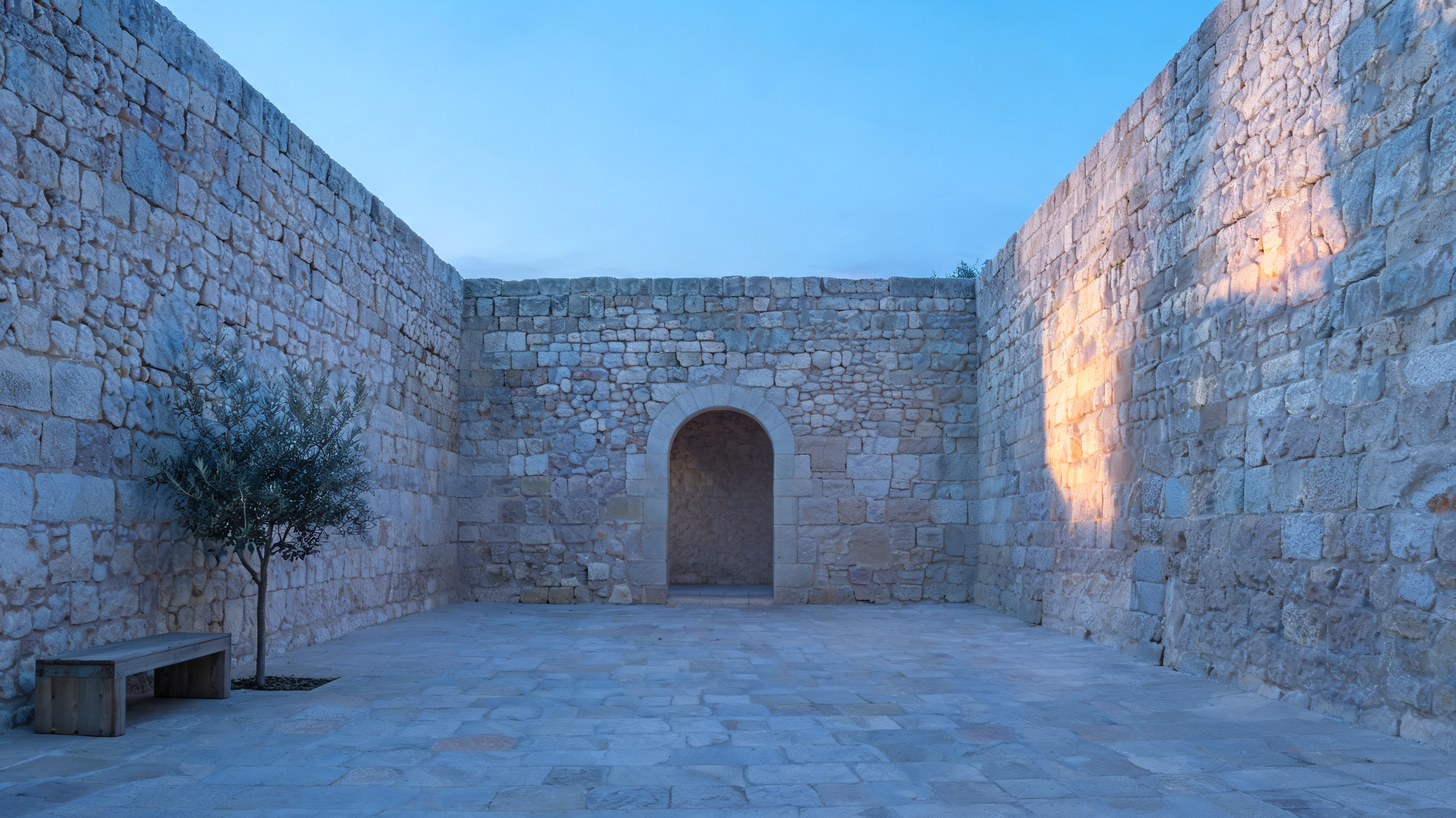 Empty Mediterranean stone courtyard at the blue hour before Fajr — pale limestone walls, a single arched doorway, soft pre-dawn light.
