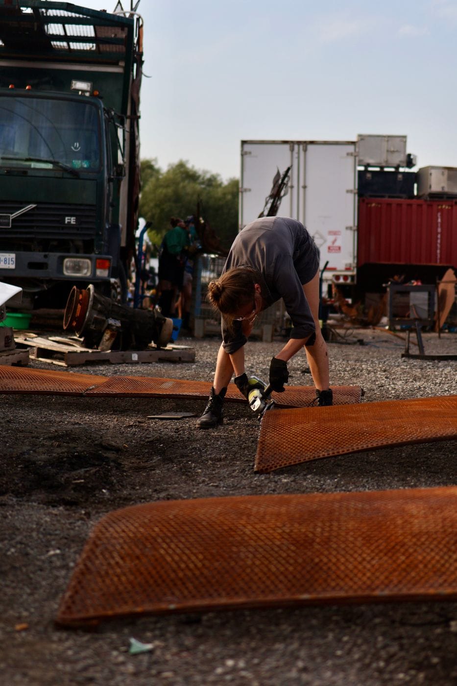 A builder bent over rusted steel panels with a grinder in a gravel yard, trucks and crew layered behind