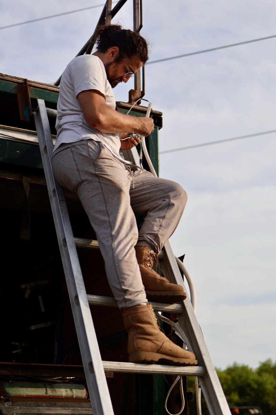 A fabricator on a ladder against the sky, wiring a cable high on a green structure