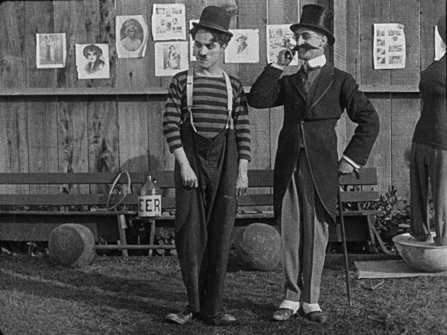 Chaplin and the villain standing next to each other. The villain has a top hat, can, and a comically large fake moustache that he's twirling.