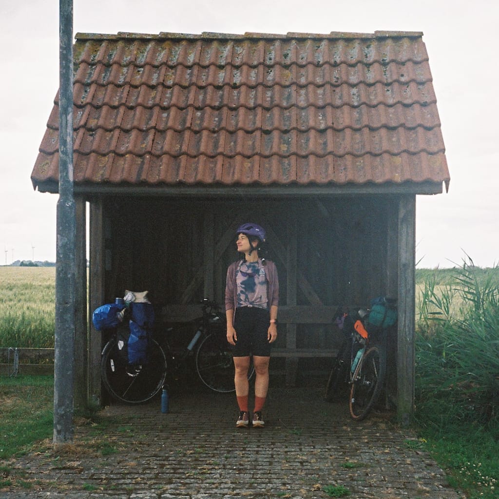 Stephanie wearing the Icon Merino T-Shirt standing under a tiny bus stop shelter next to two bikes loaded with bikepacking bags.