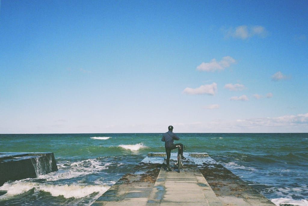Lukasz Kus looking at the Baltic Sea, hands on camera, ready to take a photo,holding his bike with one one leg propped up on the seat
