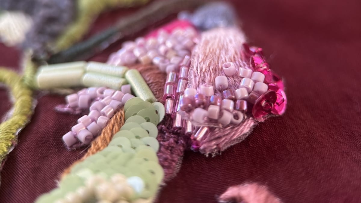 Close-up of hand embroidery on burgundy fabric, combining pink, green, yellow and ivory threads with beads and sequins.
