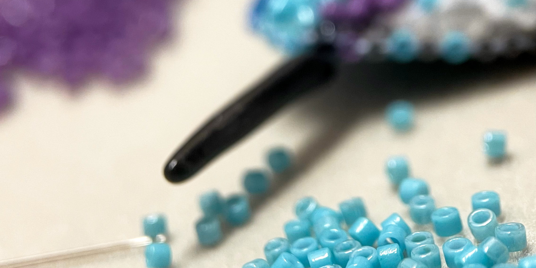 Turquoise cylinder beads on a white work surface beside a beading needle and black-handled tool, with colorful beadwork softly blurred behind.