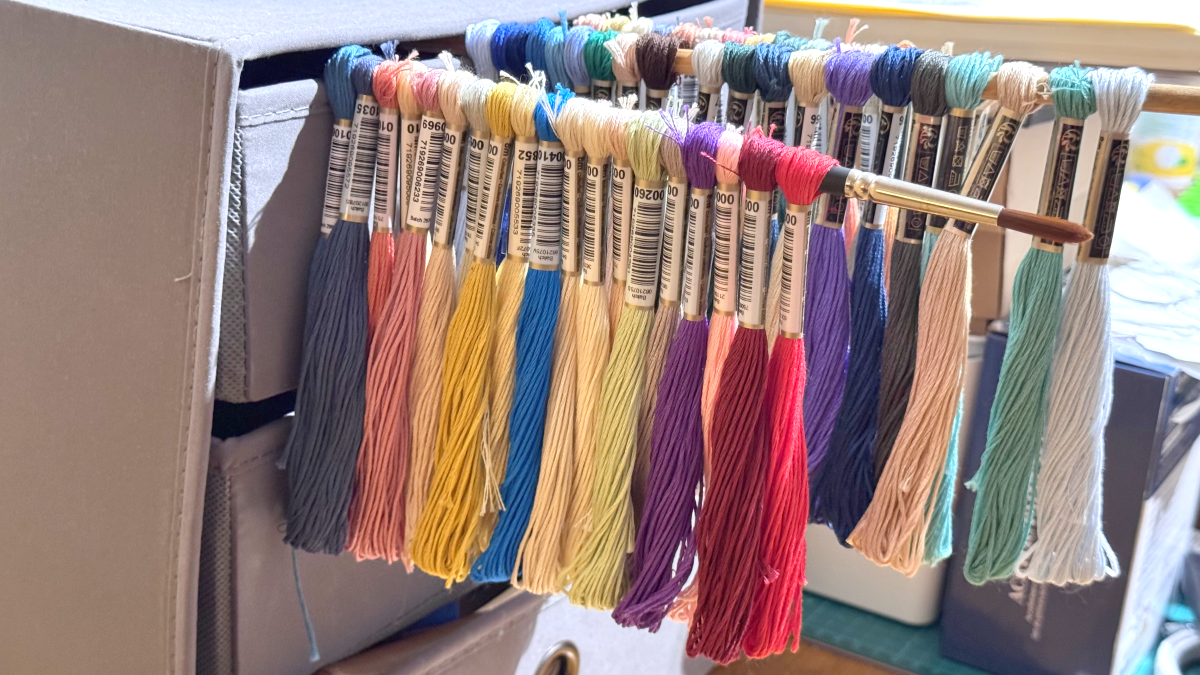 Numbered embroidery floss skeins in dark neutrals and bright blues, yellows, purples, pinks, reds, and greens hang from rods in a craft workspace.