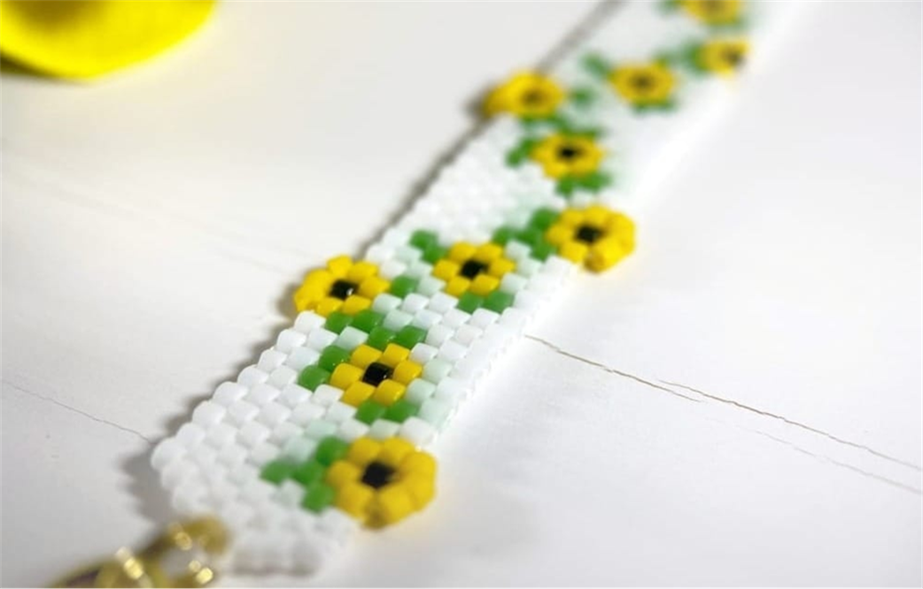 A white Miyuki Delica peyote bracelet with yellow and green sunflower motifs on a white surface.