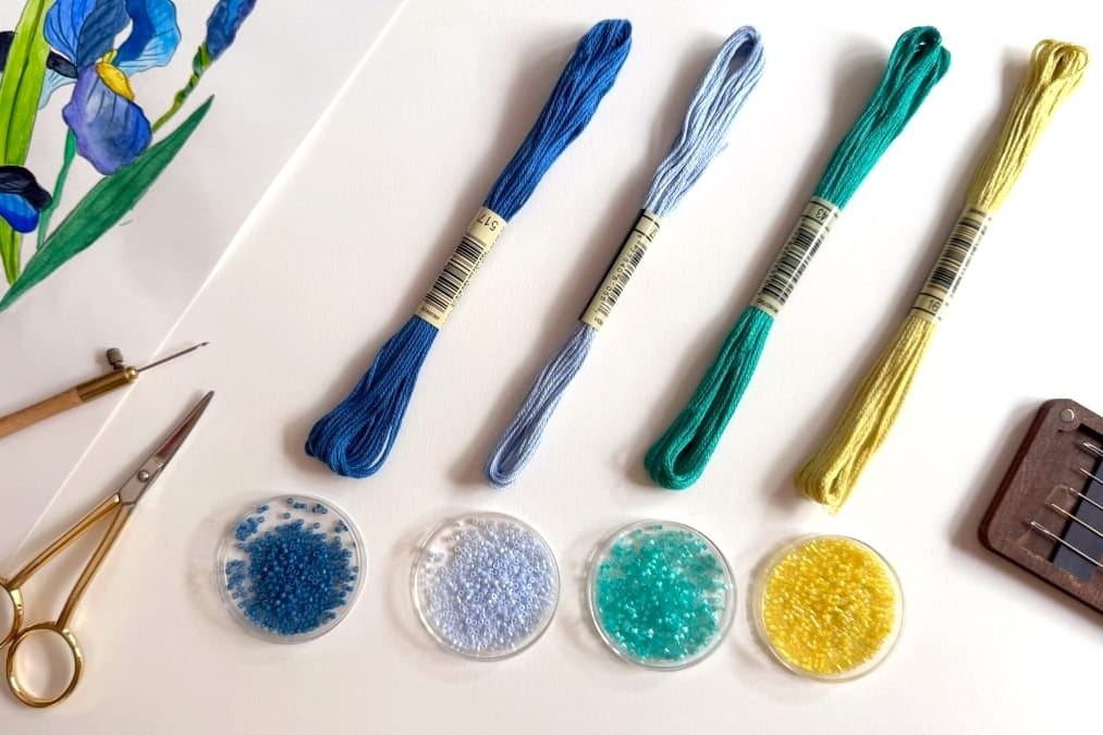 Blue, green, and yellow embroidery threads above dishes of beads, with an iris drawing and embroidery tools.