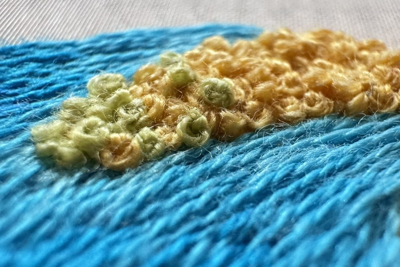 Close-up of blue embroidery stitches with raised pale green and yellow knots on white fabric.
