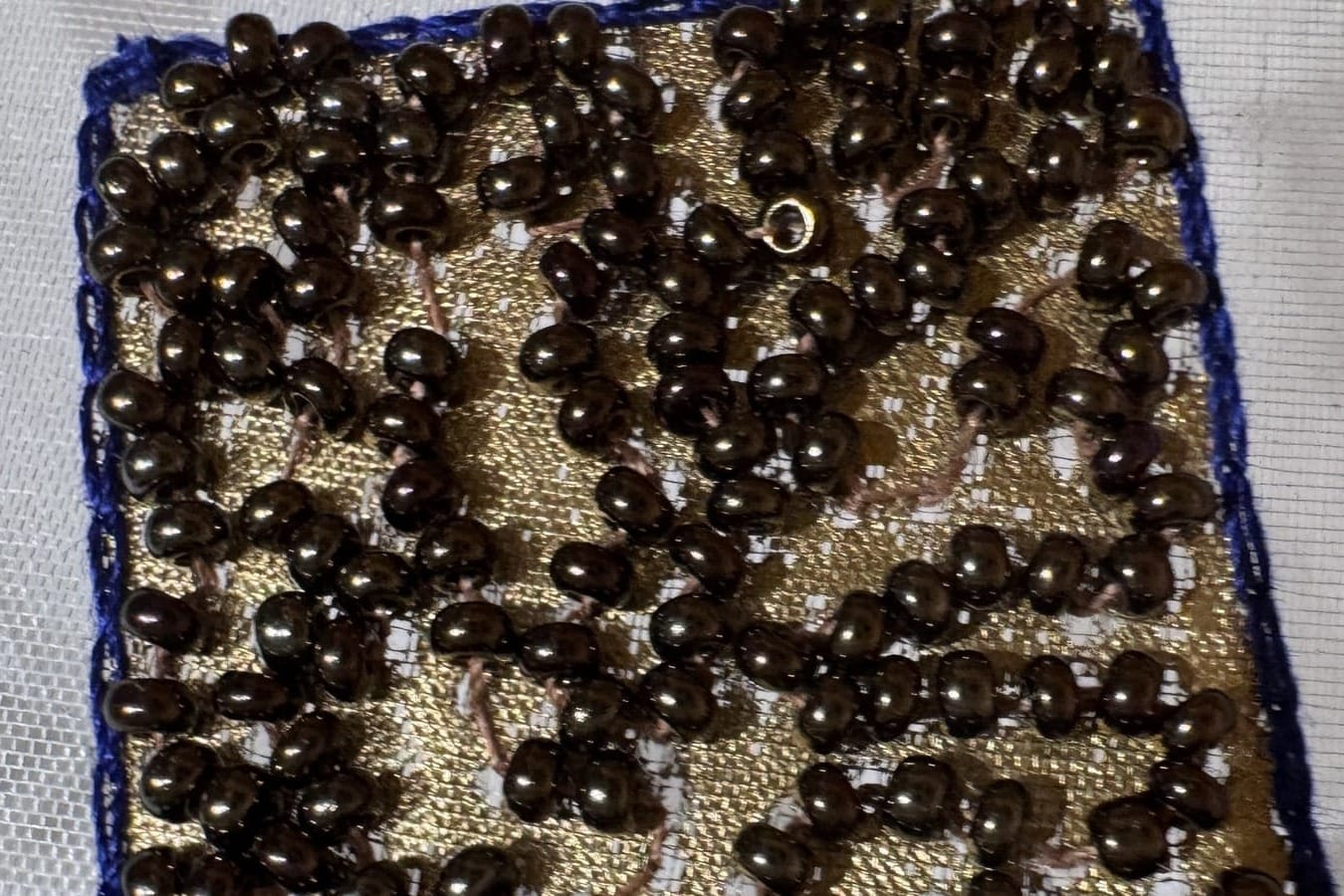 Dark seed beads stitched over a gold-colored square with a blue border on sheer fabric.