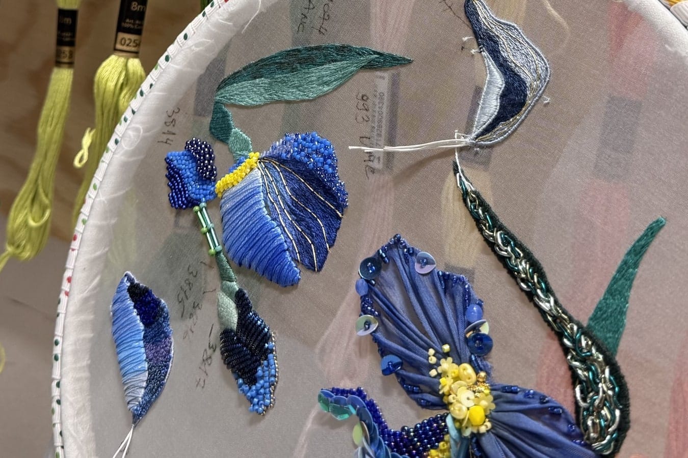 Blue iris embroidery in progress on sheer fabric in a hoop, with thread skeins hanging behind it.