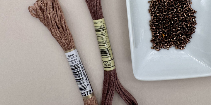 Mauve embroidery threads lies beside matte burgundy cylindrical seed beads in a white square dish on a beige surface.