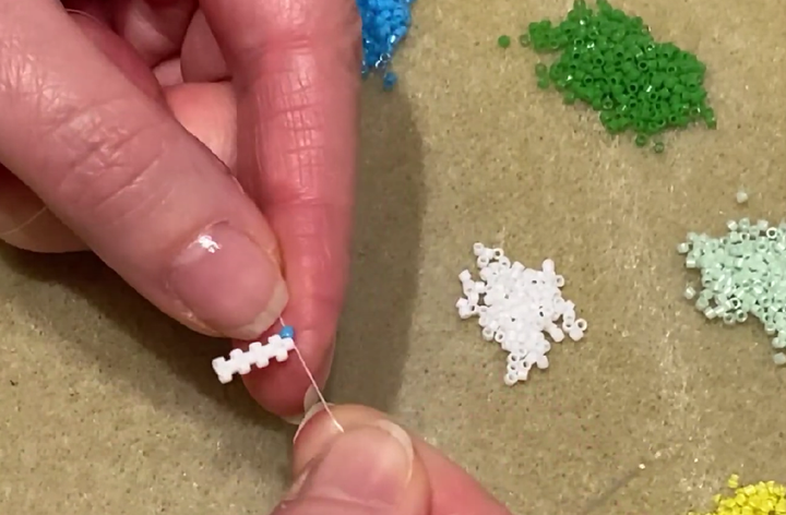Hands holding a small white peyote stitch section while a blue bead and needle begin the next row.
