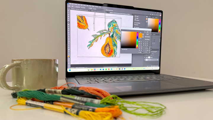 A laptop displays a colorful botanical design and an open color picker, with green, orange, yellow, and black embroidery floss beside a ceramic mug