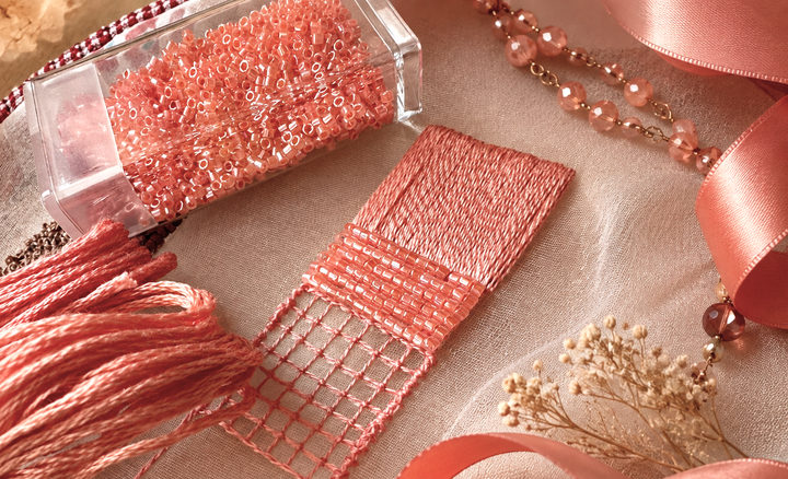 Coral-pink cylindrical seed beads in a clear box beside matching embroidery floss woven through a small open-grid sampler, with ribbon and jewelry on beige fabric