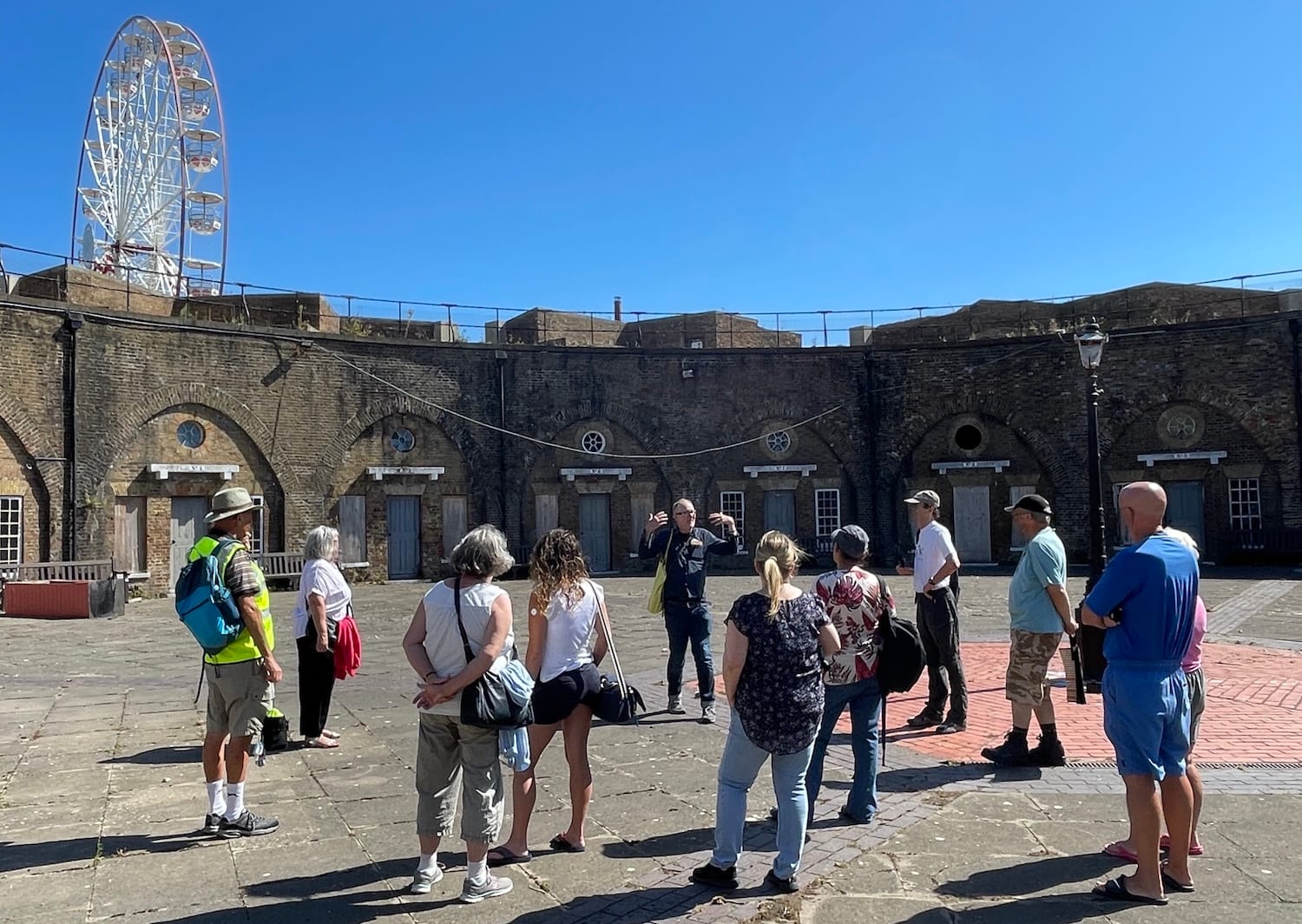 A man talks to a dozen people standing inside a large fortress against a blue sky
