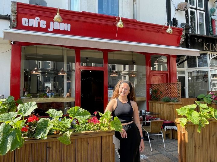 A young woman smiling by summer flowers outside a red-painted cafe exterior