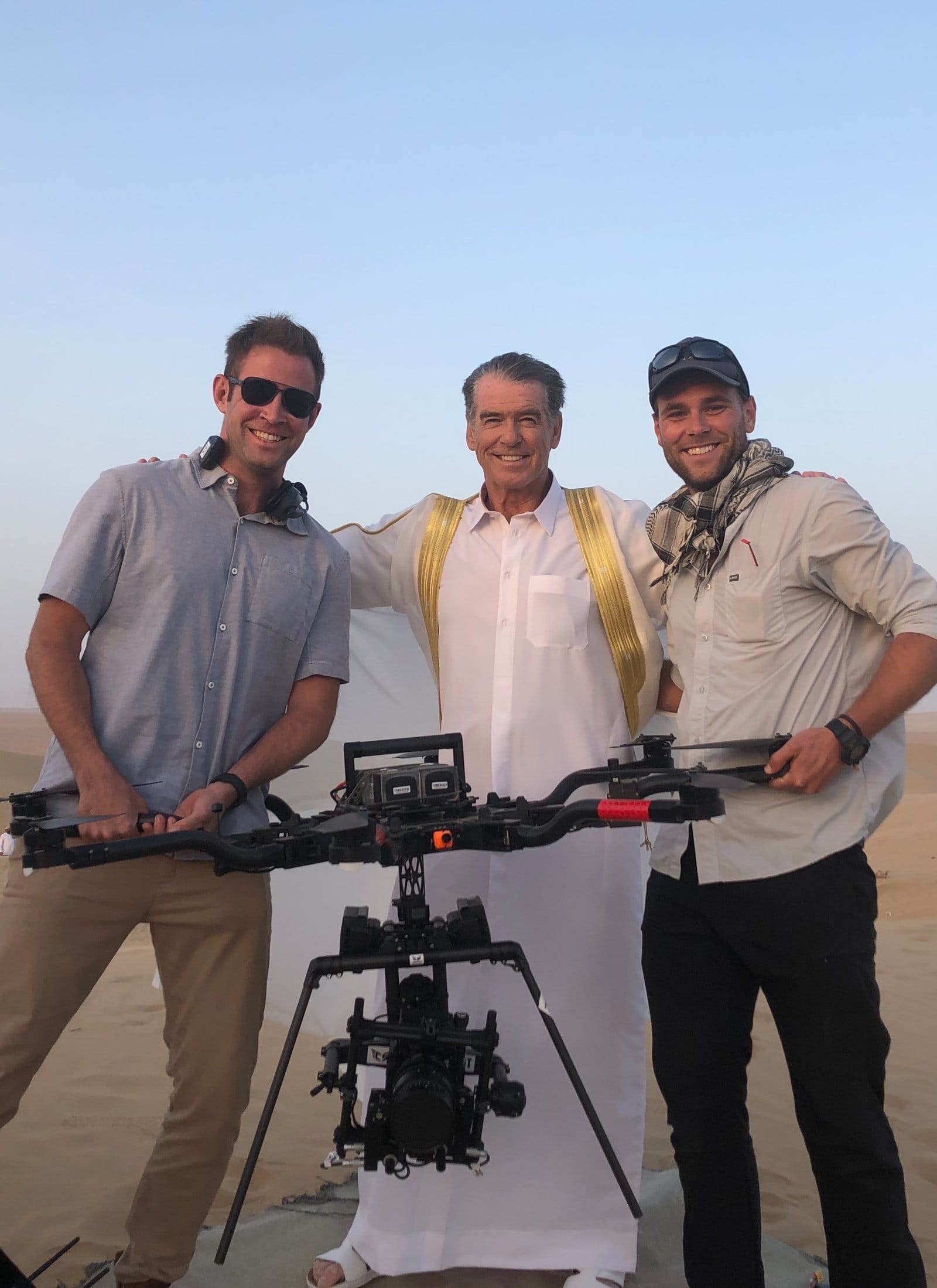 Rob, his flight partner Pat, and Pierce Brosnan standing behind a large aerial cinematography drone rig in a desert setting