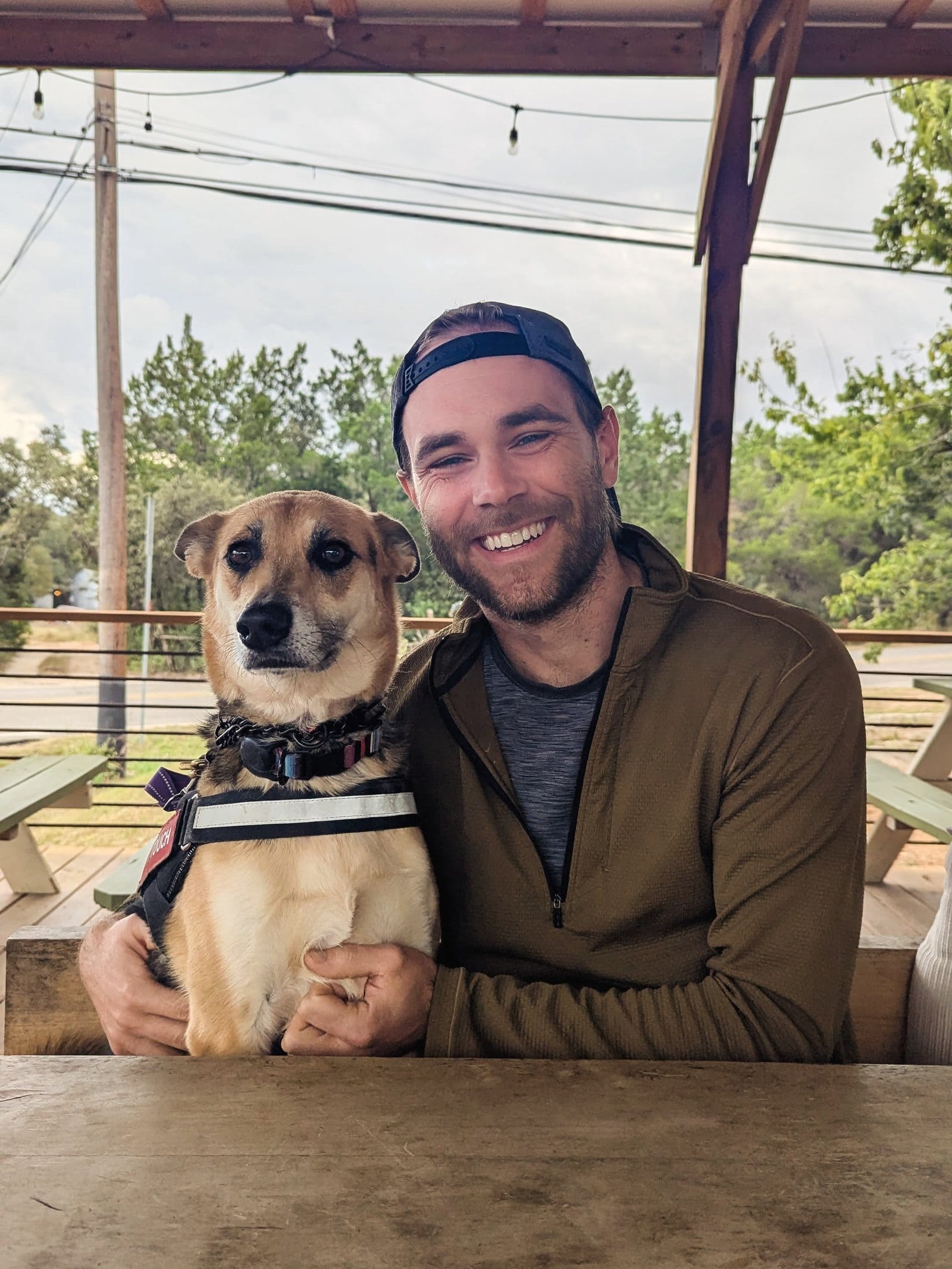 Rob smiling next to his Belgian Malinois Chai, who is wearing a harness and sitting at an outdoor cafe table
