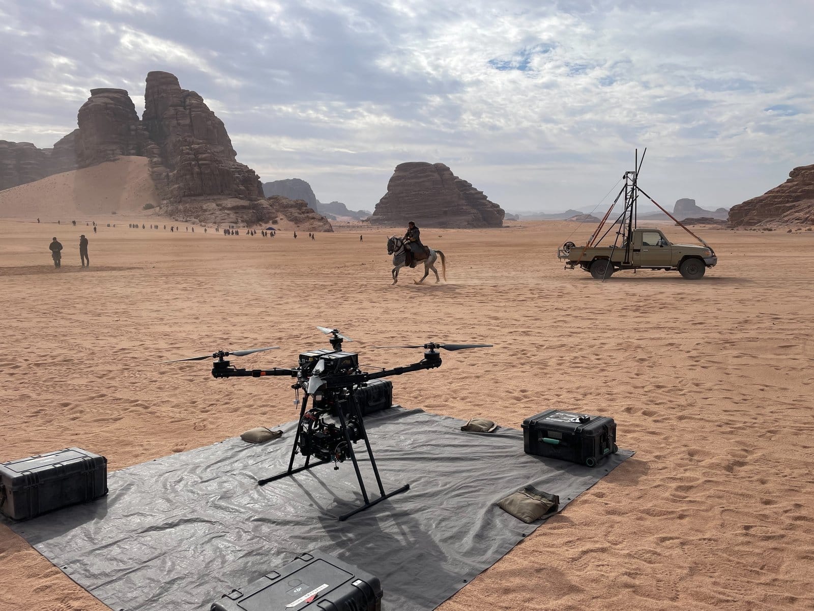 A heavy-duty aerial cinematography drone on its landing pad in the foreground, a horseback rider crossing the desert behind, and red sandstone mountains of NEOM in the distance
