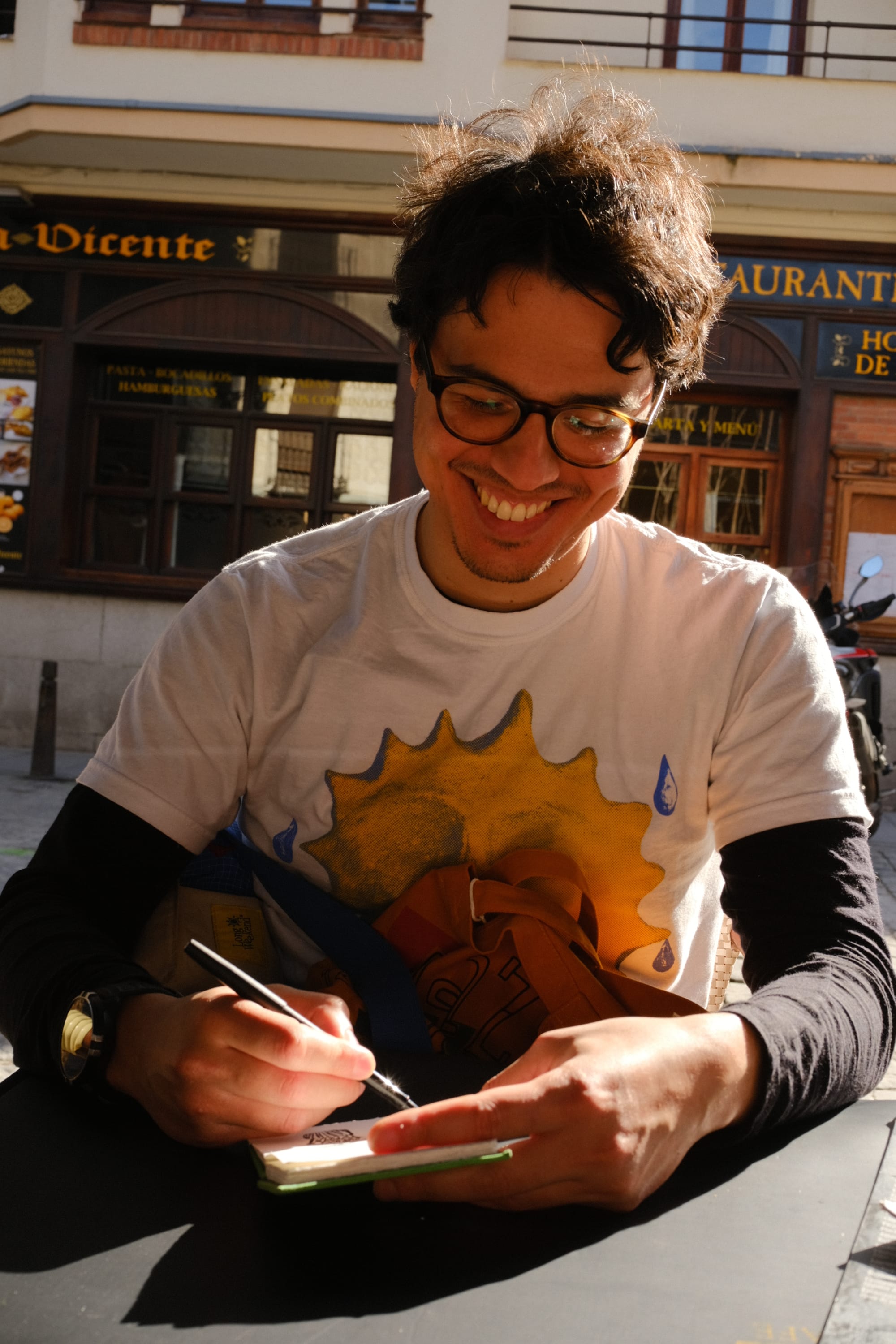 Mateus sits at an outdoor café table, smiling as he looks down into a notebook, mid sketching with a pen.