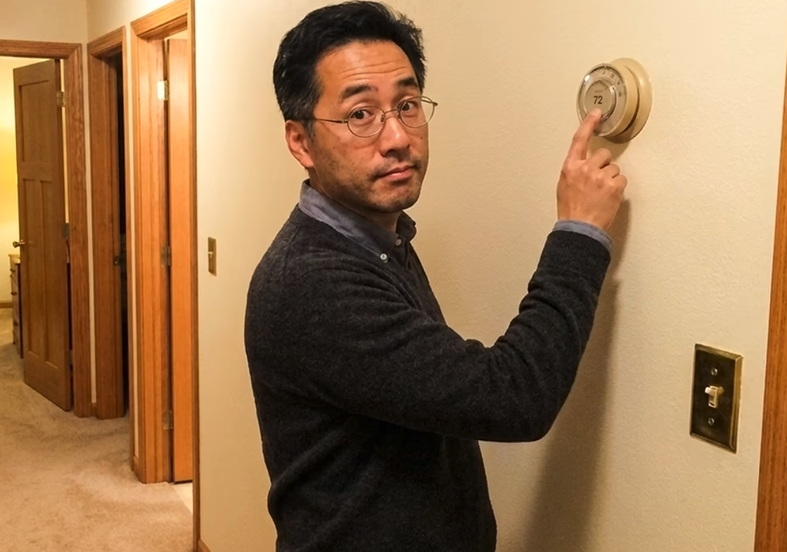 "Ben" stands in the middle of an apartment hallway, pointing a finger at a thermostat and staring at the camera with a mildly-interested expression