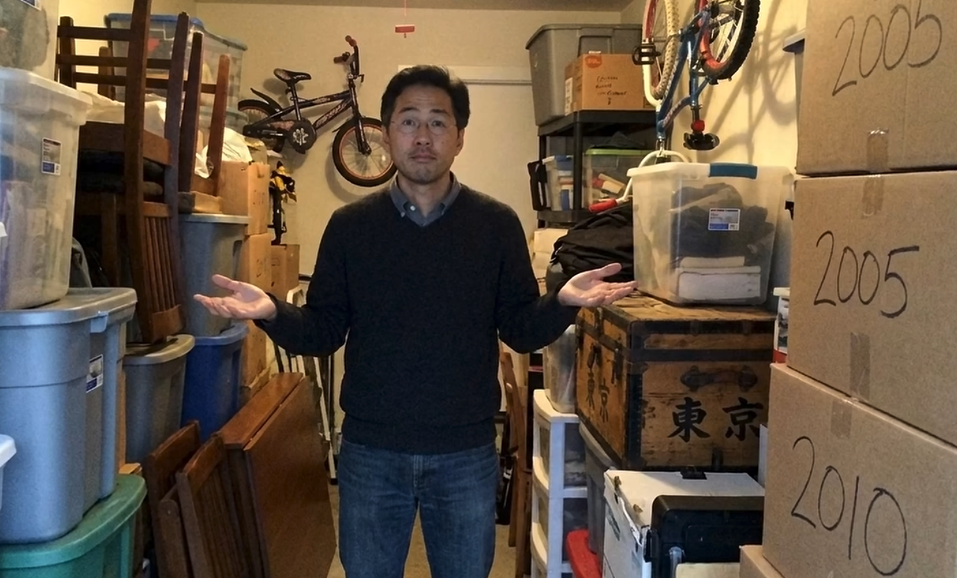 "Ben" stands in front of many boxes and other household objects like chairs and shelves. He stares at the camera, shrugging his arms.