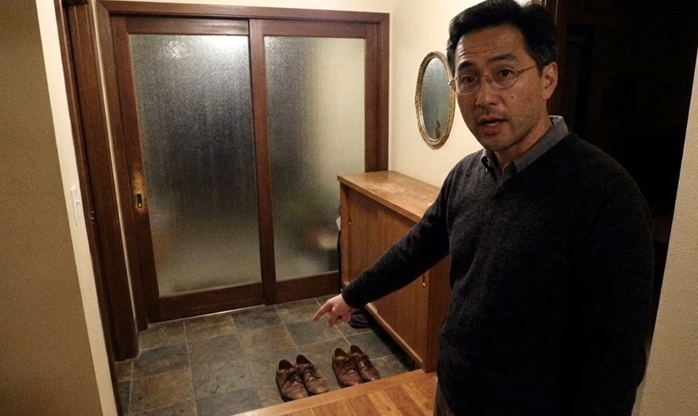 "Ben" stands near a house entrance, pointing at two pairs of shoes near the door. He stares at the camera, with his mouth slightly open. The entrance is placed at a slightly lower elevation from the rest of the house.