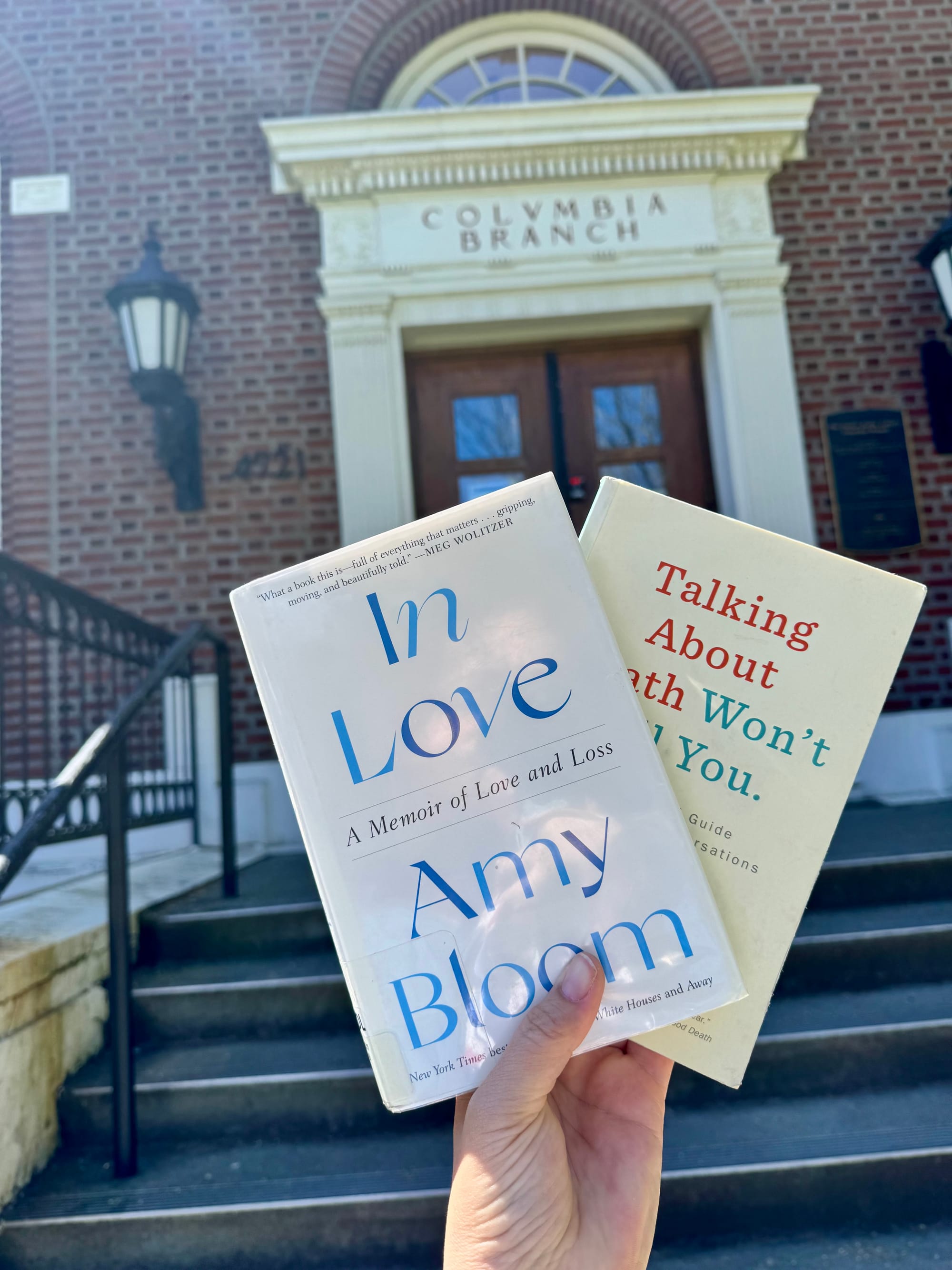 A picture of a hand holding In Love by Amy Bloom, and Talking About Death Won't Kill You