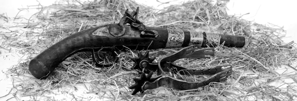 A flintlock pistol and a set of spurs lie in scattered hay. black and white image.