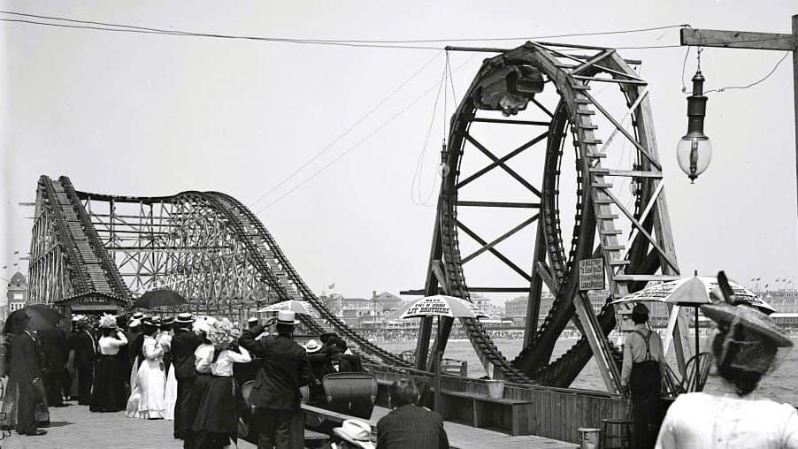 Black and white glass plate photograph of a wooden looping roller coaster in Atlantic City circa 1901. People watch on the sides and a car is at the apex of the loop.