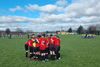 Kids in red shirts huddle in a circle on a green sports pitch, under a blue sky